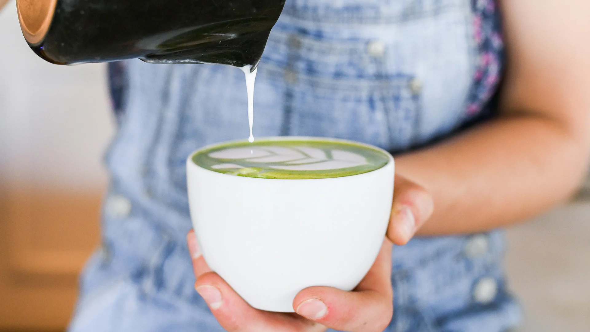 A person holding a white cup filled with matcha latte, pouring milk to create latte art.