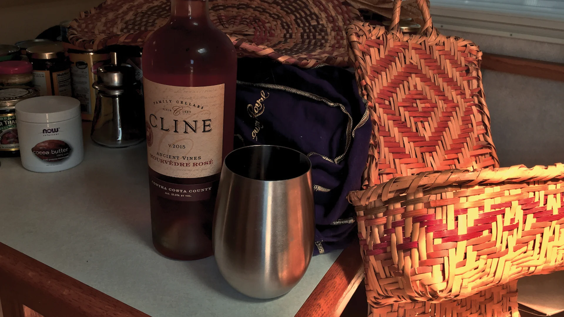 A bottle of Cline rosé wine stands next to a metal wine tumbler on a kitchen counter. Nearby are woven baskets, jars, and a dark cloth bag in a warmly lit setting.
