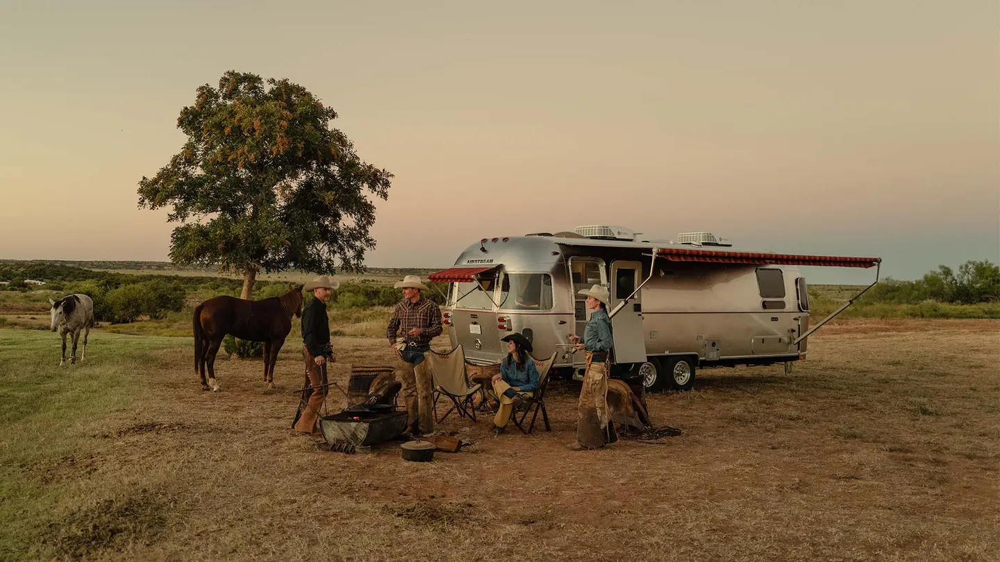 Four people and a dog gather around a campfire near a silver camper and horses on a grassy plain at sunset.