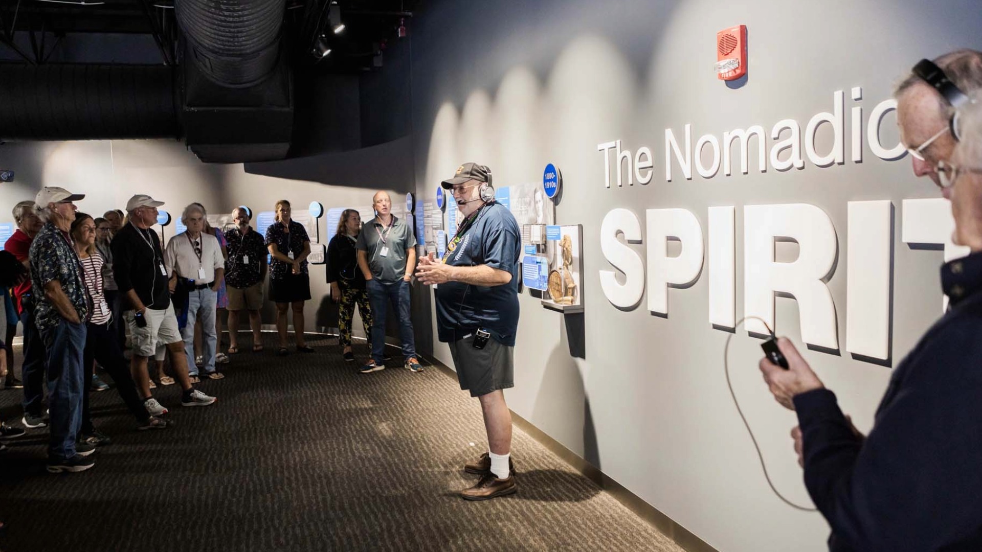 A group of people listens to a tour guide in a museum next to a display titled "The Nomadic Spirit." Some wear headphones and casual attire.