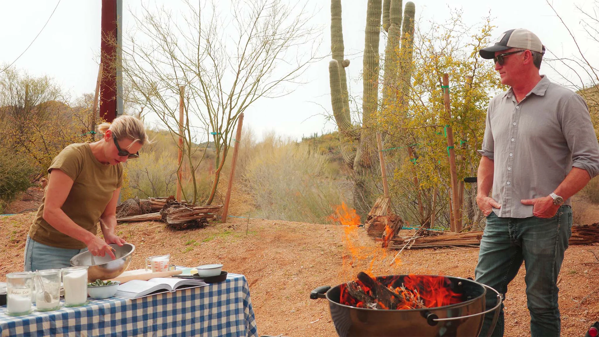 A woman prepares food at a table with bowls and a book. A man stands nearby. A fire burns in a pit. Desert plants are visible in the background.
