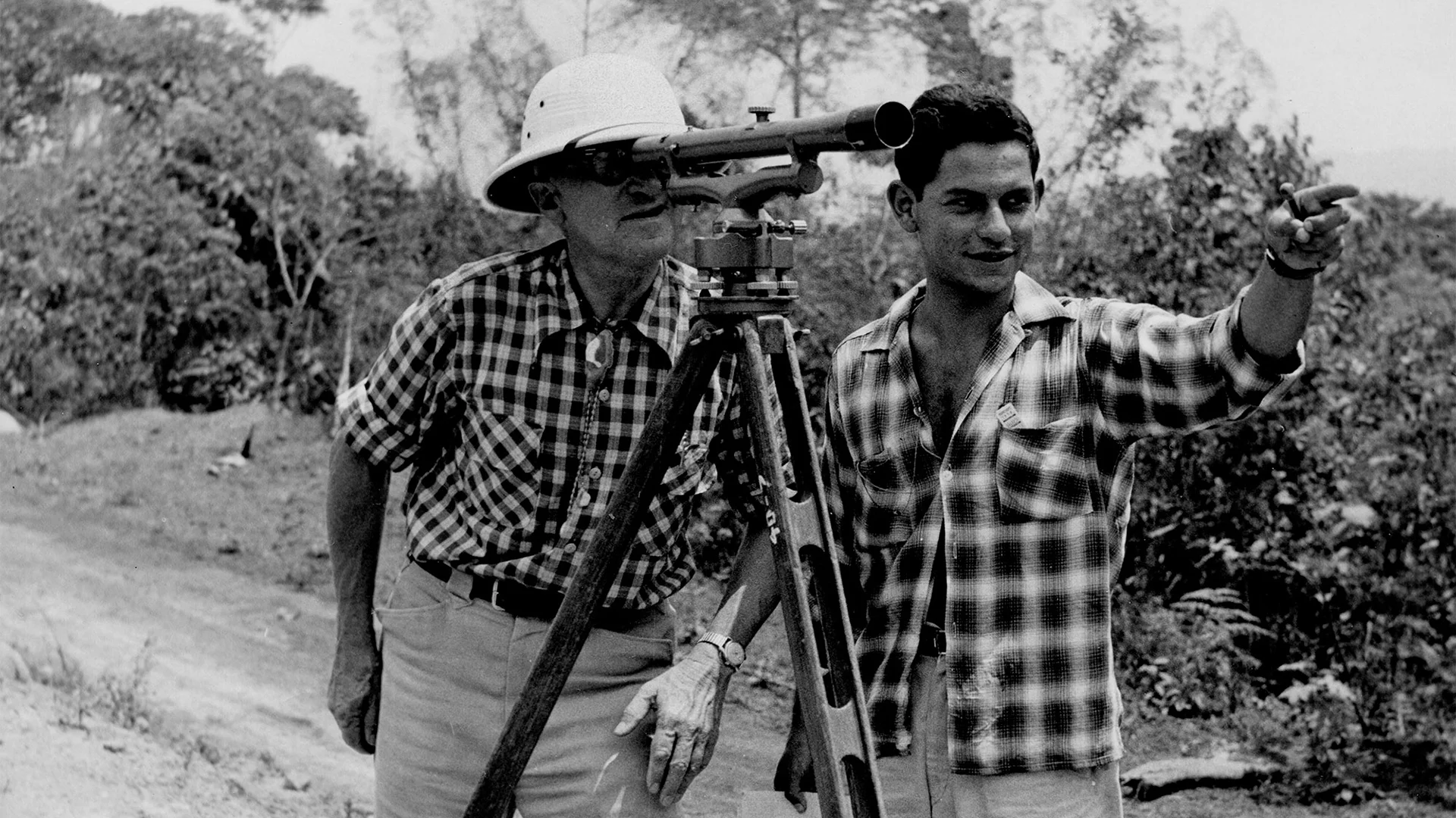 Two men in checkered shirts survey a landscape using a tripod-mounted telescope. One points ahead while the other looks through the lens.