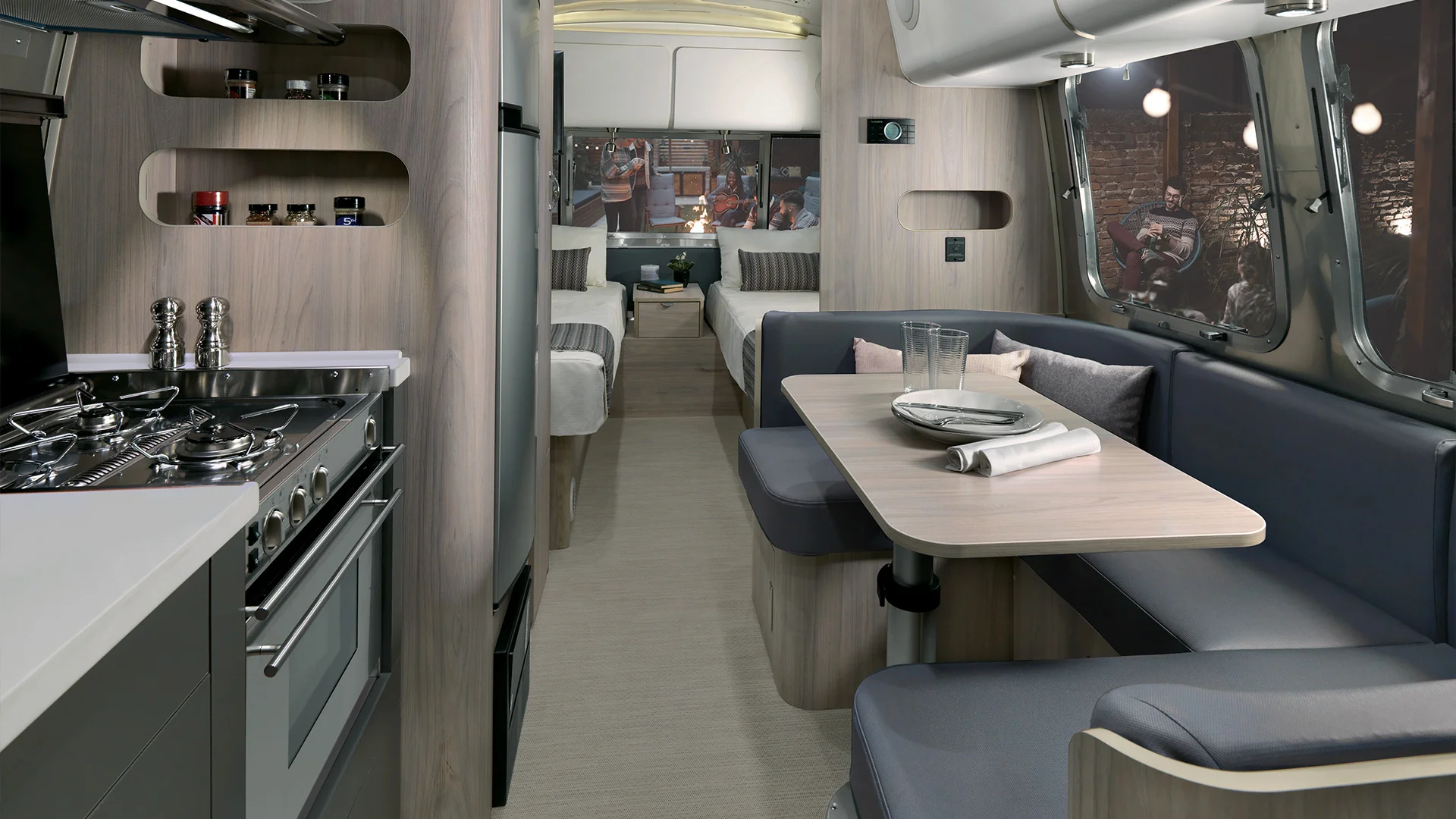 Modern RV interior with a kitchen, dining nook, and twin beds. A large window offers a view of people outdoors.