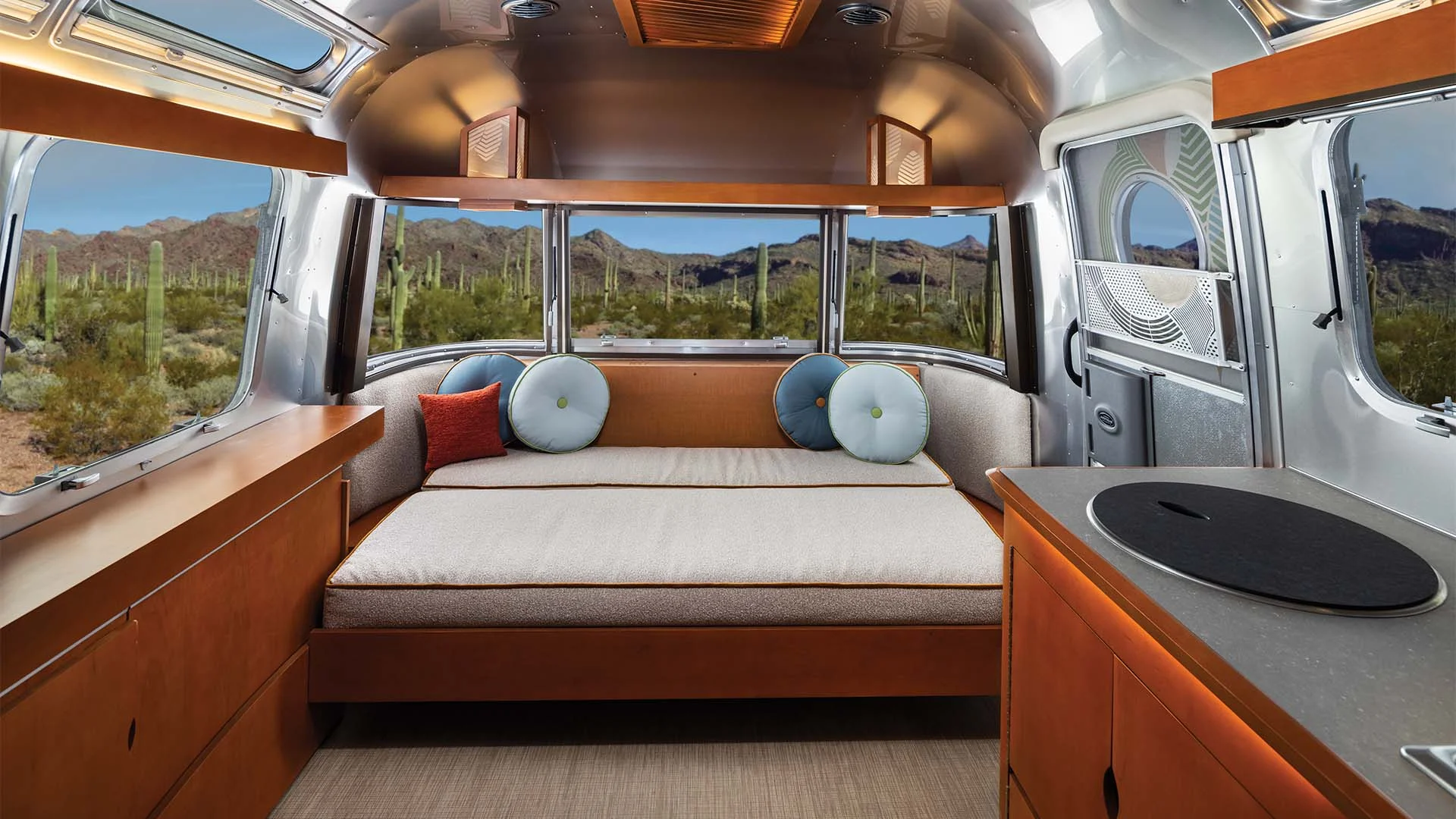 Interior of an Airstream trailer with a bed, decorative pillows, wooden cabinets, and large windows overlooking a desert landscape with cacti.