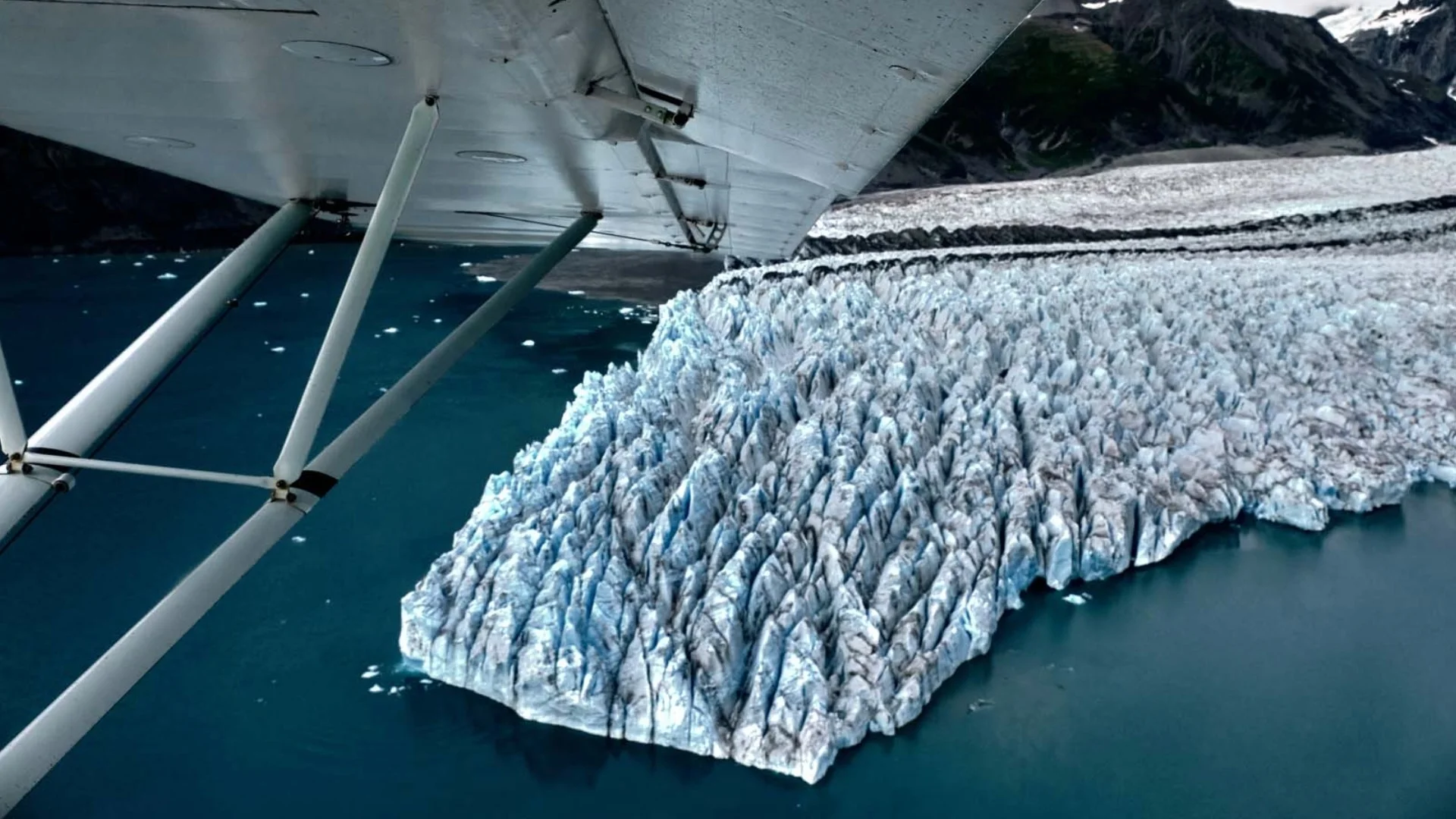 Aerial view of a glacier with jagged ice formations meeting the blue ocean, seen from beneath an airplane wing.