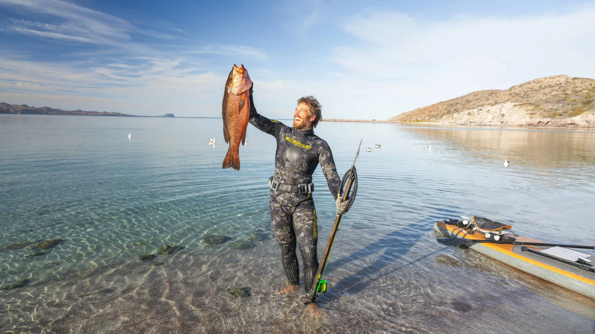 A man in a wetsuit joyfully holds up a large fish by the shore, standing beside a kayak on a clear, sunny day.