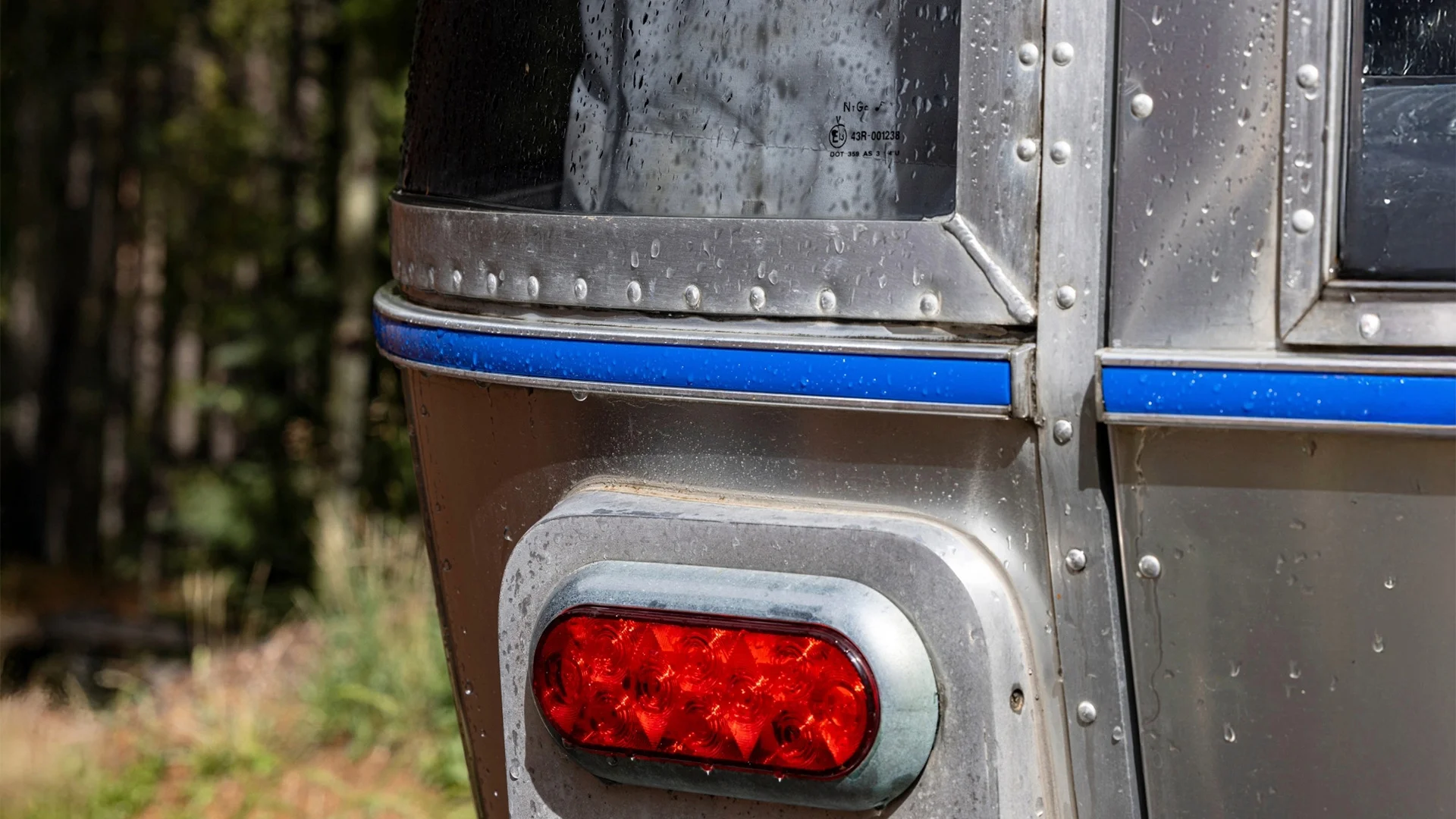 Close-up of a vintage Airstream trailer corner, showing red tail light, riveted silver metal exterior, and raindrops. Trees blurred in the background.