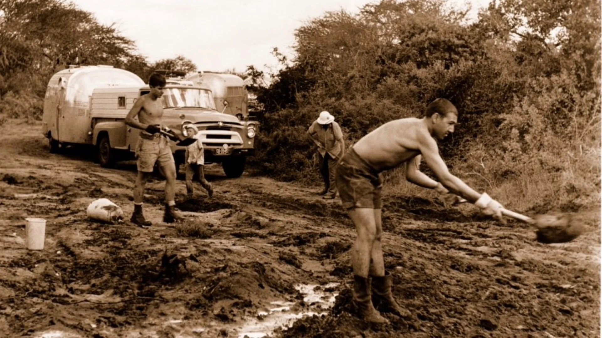 Men in shorts and boots work on a muddy road with shovels, near vintage vehicles and trees, under an overcast sky.