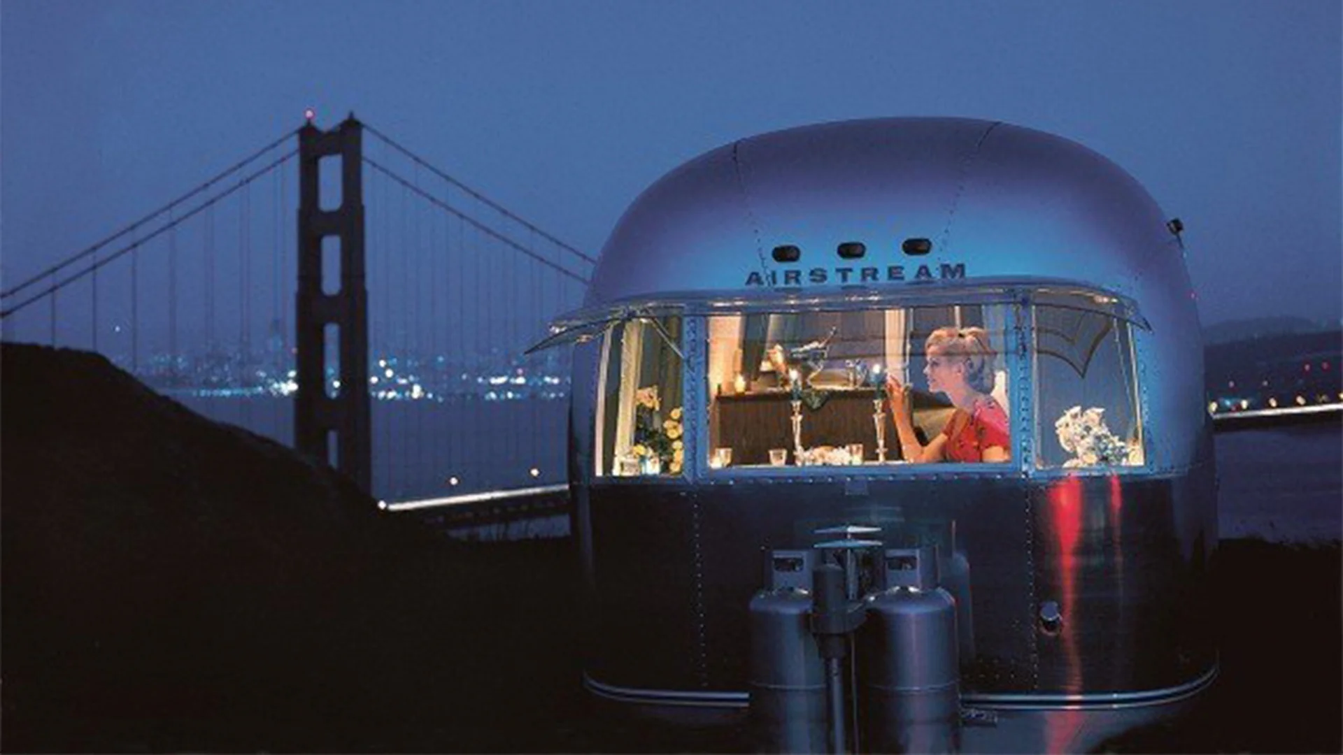 Airstream trailer at night with a view of a lit-up bridge. A person inside is seated at a table with decorative items.