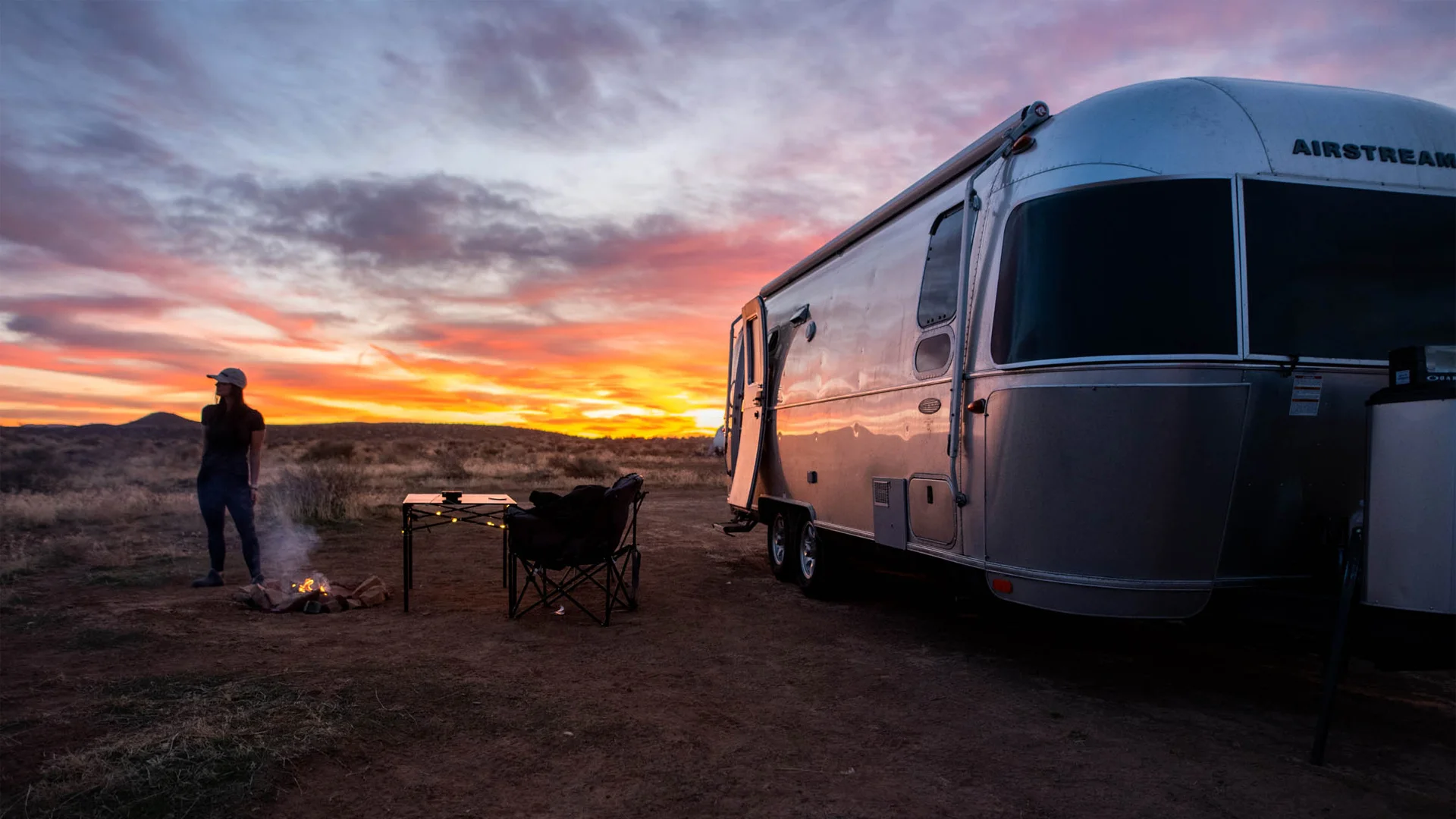 A person stands near a campfire and folding chairs beside a silver camper trailer on dry grassland at sunset, with colorful clouds in the sky.