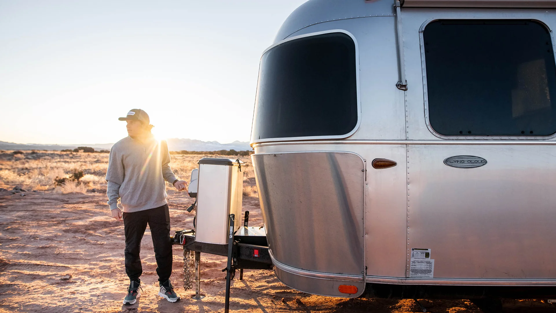 A person stands beside a modern silver trailer at sunrise in a desert landscape, wearing casual clothes and a hat.