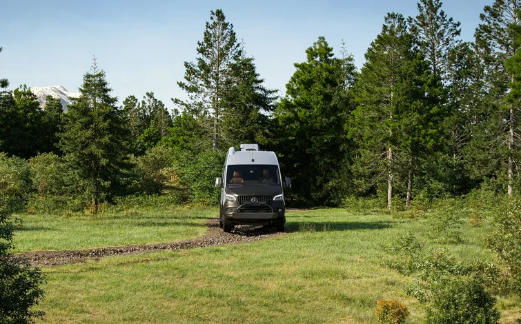 A camper van driving on a narrow dirt path in a lush forest with tall pine trees and a mountain partially visible in the background.