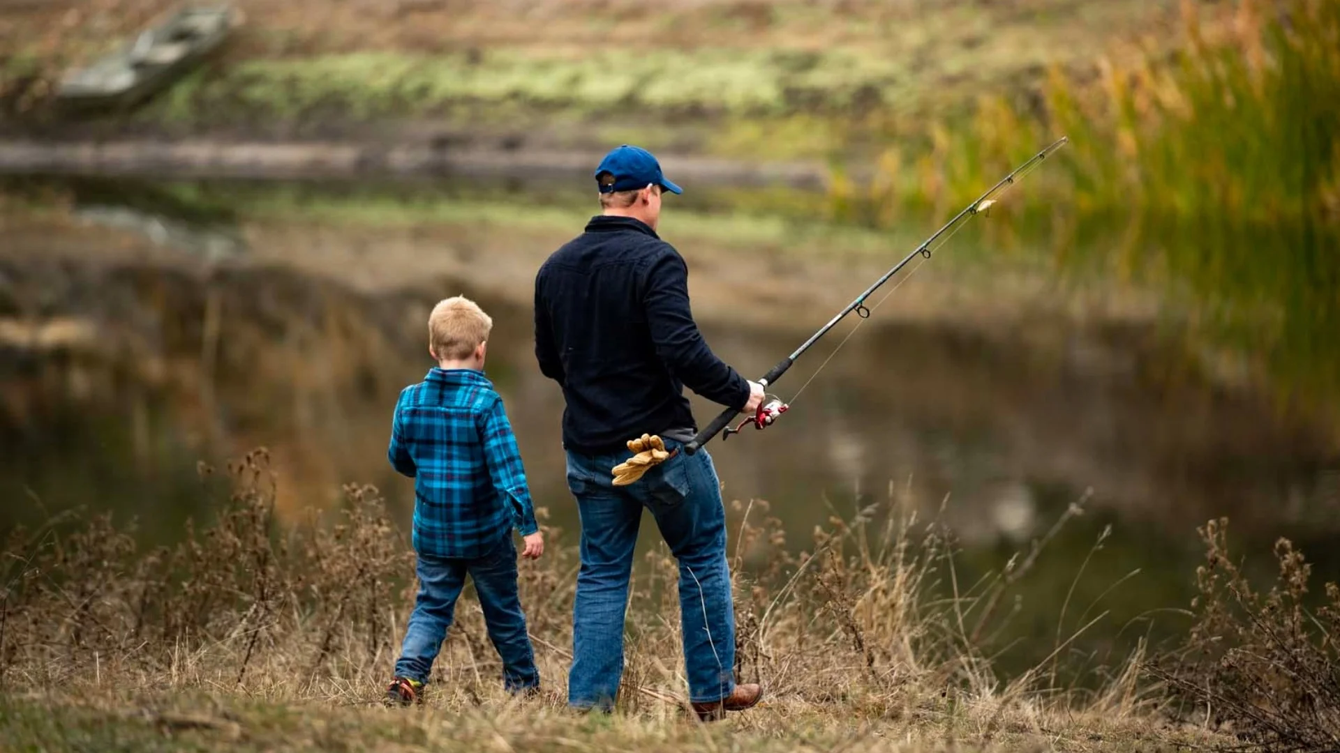 A man and a child stand on a grassy bank, holding fishing rods, gazing at a calm pond surrounded by greenery.
