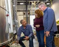 Three people in a workshop inspect the exterior of a silver trailer, with tools and boxes nearby, and a ladder in the background.