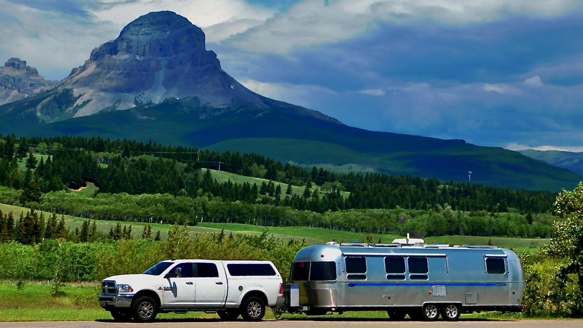 A white pickup truck towing a silver Airstream trailer is parked on a road with lush green fields and forests, set against a dramatic mountain with a rocky peak under a partly cloudy sky.