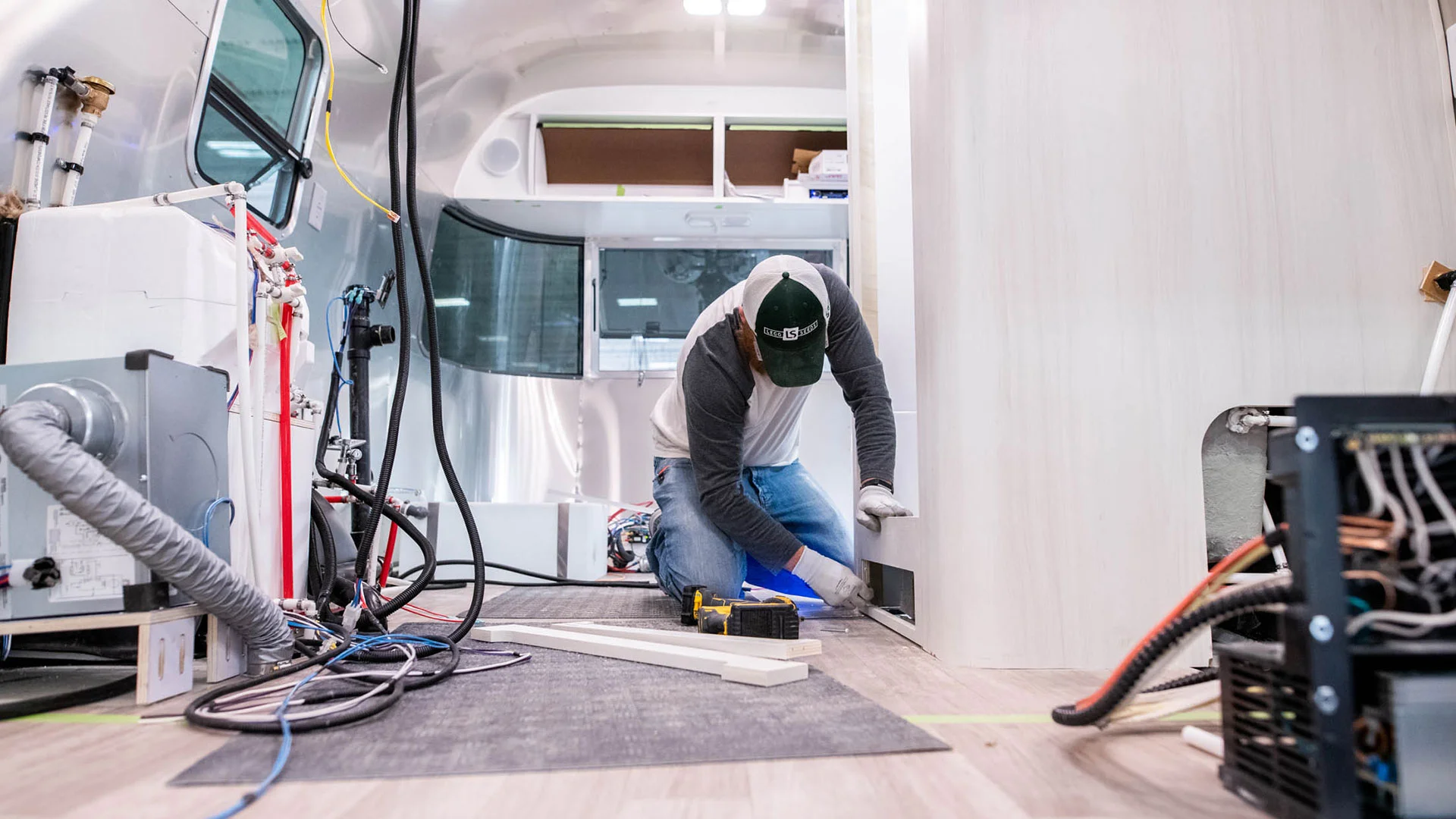 Person in a cap and gloves installing interior components in a modern RV, surrounded by tools and wiring, under bright lighting.