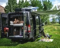 A person relaxes in bed inside a camper van parked on grass with its back doors open. An awning is extended, and folding chairs and a small table are set up outside, surrounded by trees and mountains.