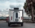 A shiny Airstream trailer hitched to a red vehicle is parked by a beach, with palm trees and a cloudy sky in the background.