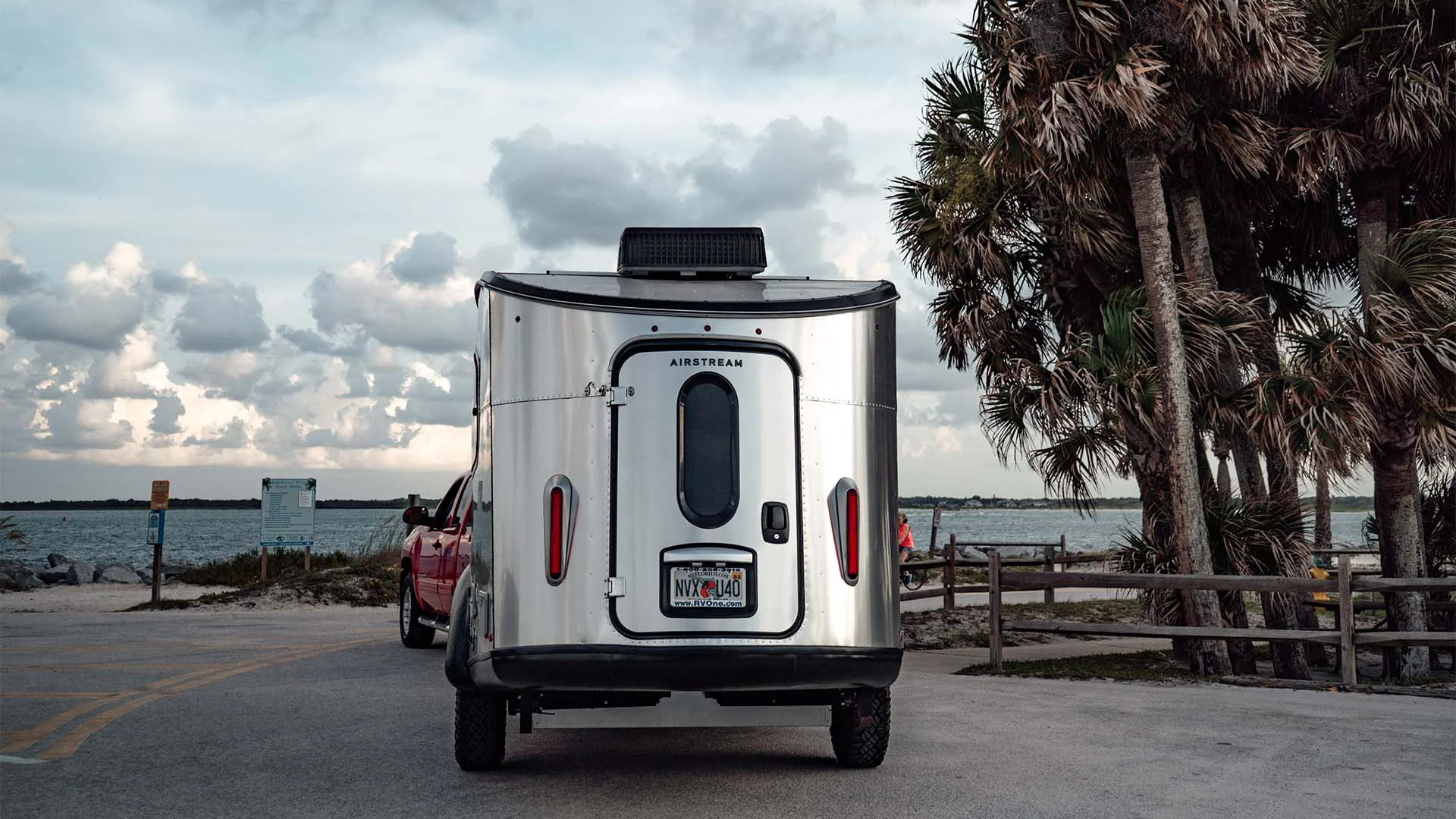 A shiny Airstream trailer hitched to a red vehicle is parked by a beach, with palm trees and a cloudy sky in the background.