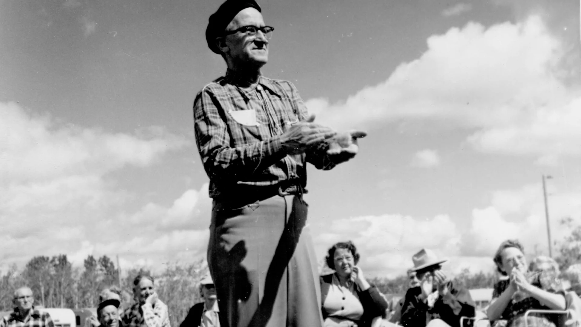 An older man wearing glasses, a beret, and plaid shirt stands outdoors clapping, while a group of people seated behind him smile and watch. The sky is partly cloudy and the scene appears festive.