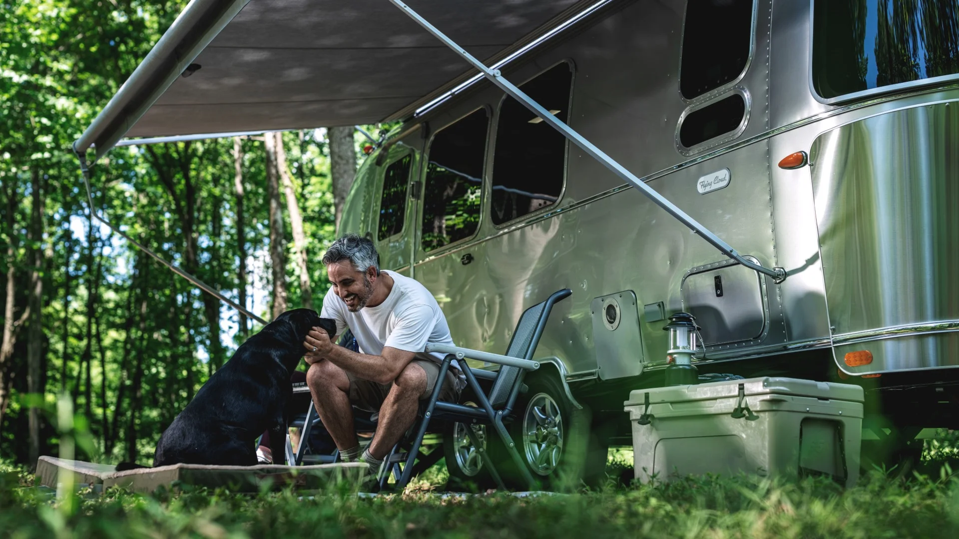 A man petting a black dog under the awning of a silver Airstream trailer in a wooded area, with a lantern and cooler nearby.