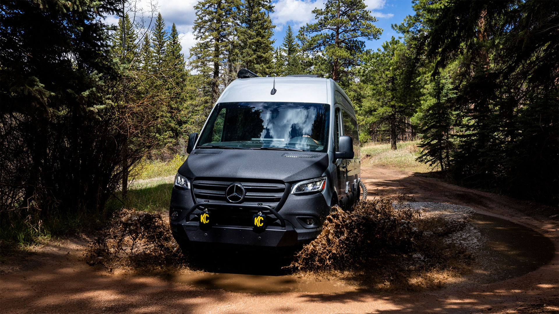 A black Mercedes van driving through a muddy forest trail, splashing water amid tall trees under a partly cloudy sky.