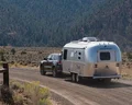 SUV towing an Airstream trailer on a scenic dirt road, surrounded by dry grass and distant forested hills.