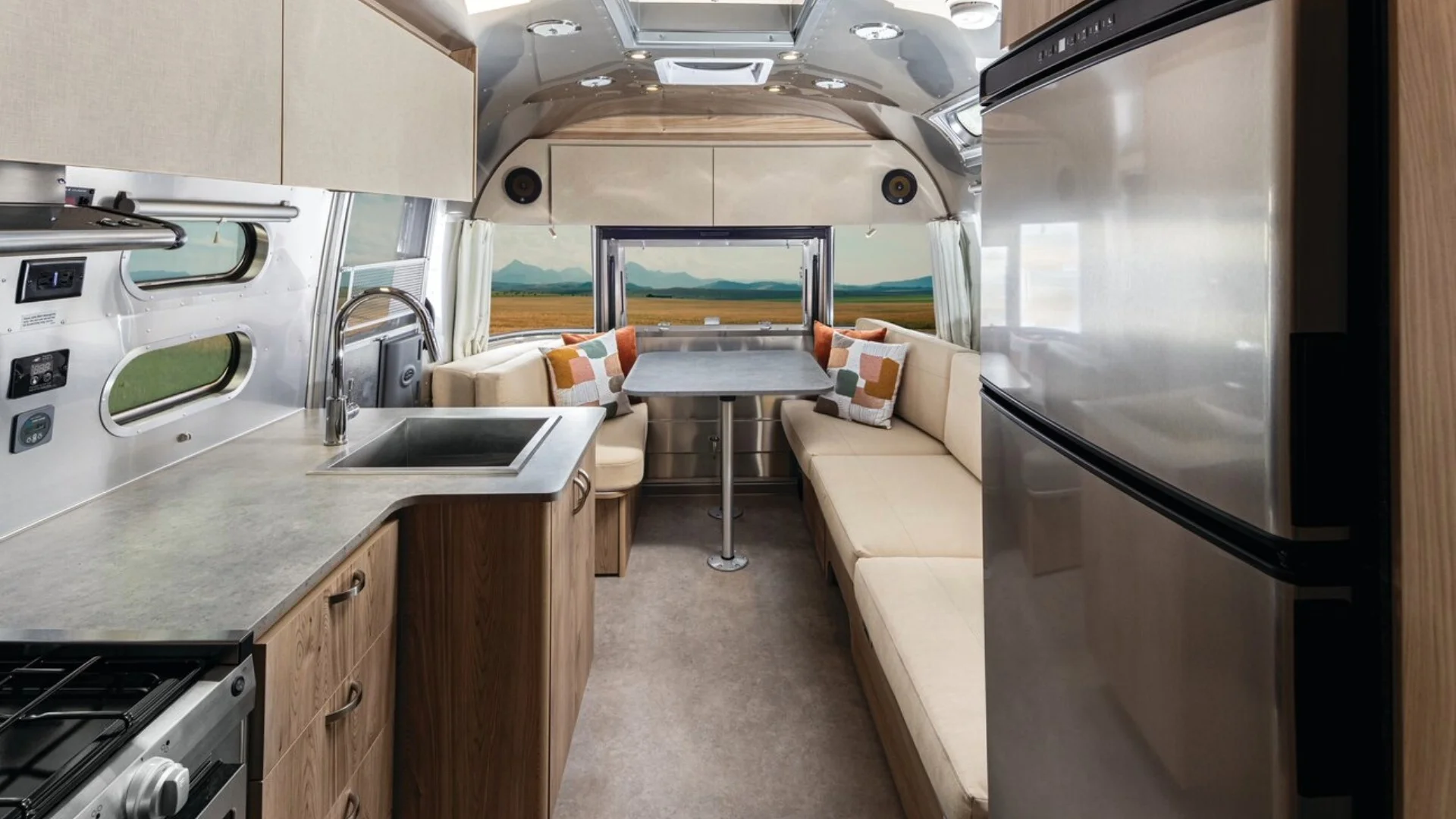 Modern RV interior with a kitchen, stainless steel fridge, and a cozy dining area with a table and beige seating, viewed through large windows.
