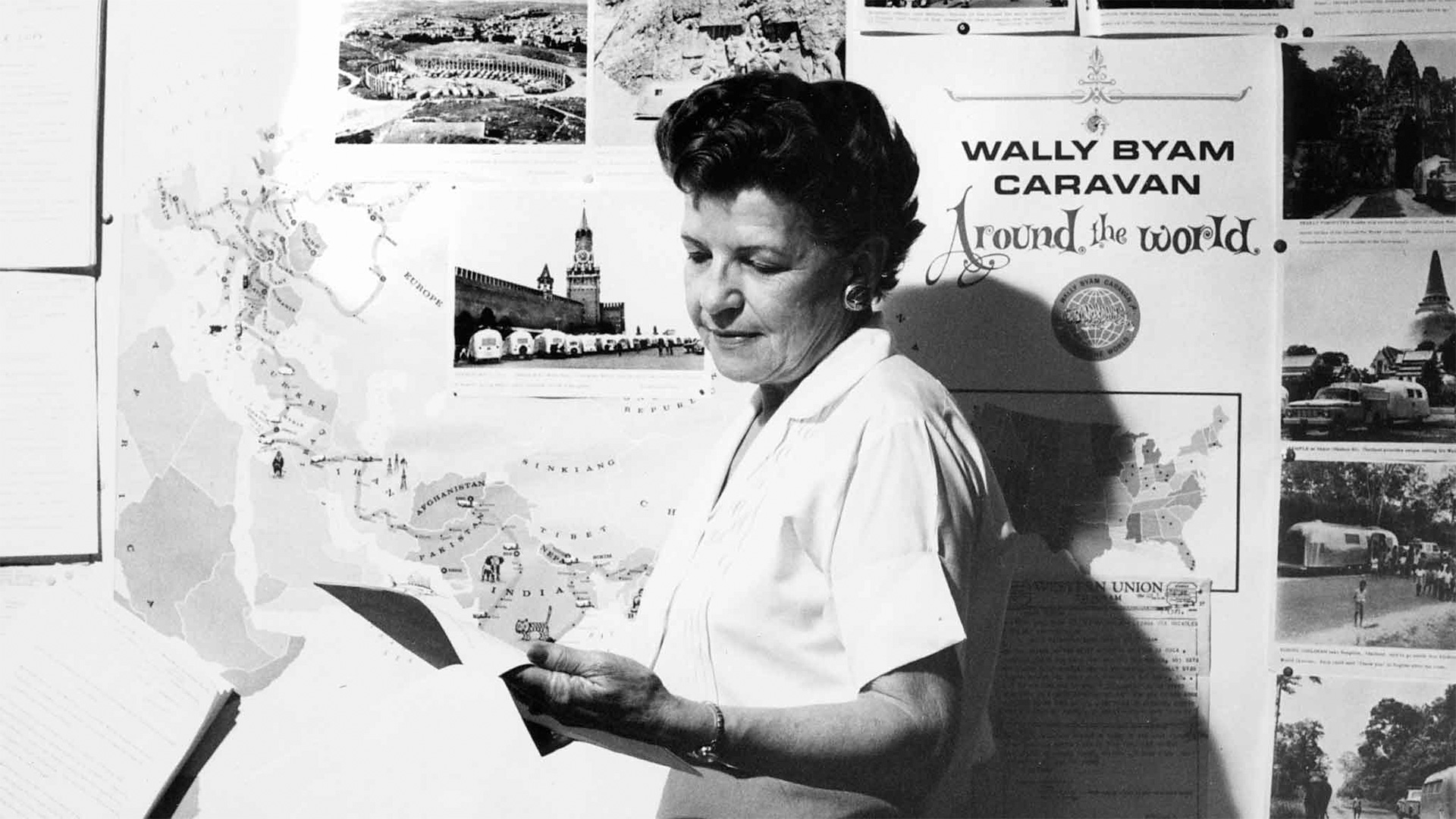 A woman examines a document in front of a wall filled with maps and posters related to Wally Byam caravans.