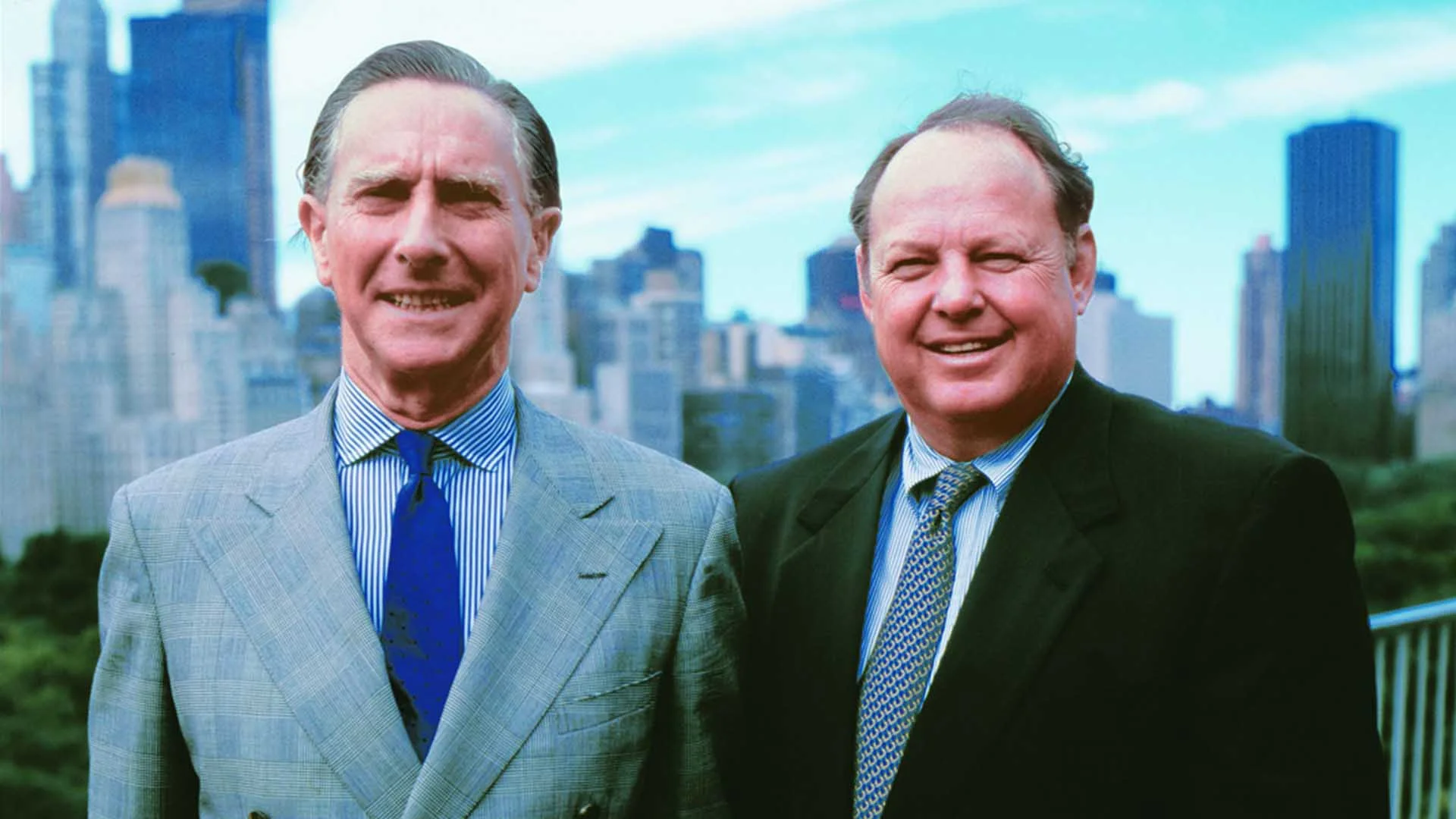 Two men in suits stand outdoors, smiling, with a city skyline and green trees visible in the background under a blue sky.