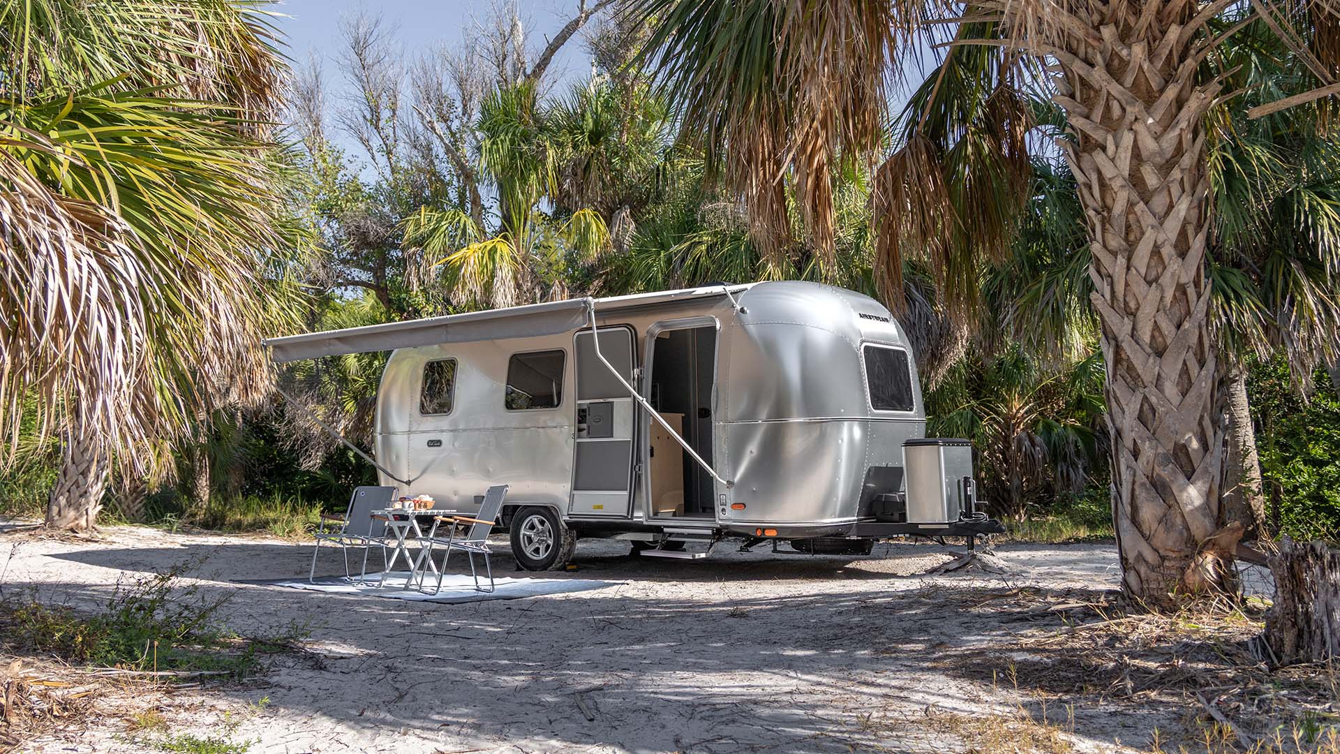 A sleek, silver camper trailer with an extended awning is set up in a tropical campsite with tall palm trees and sandy ground.