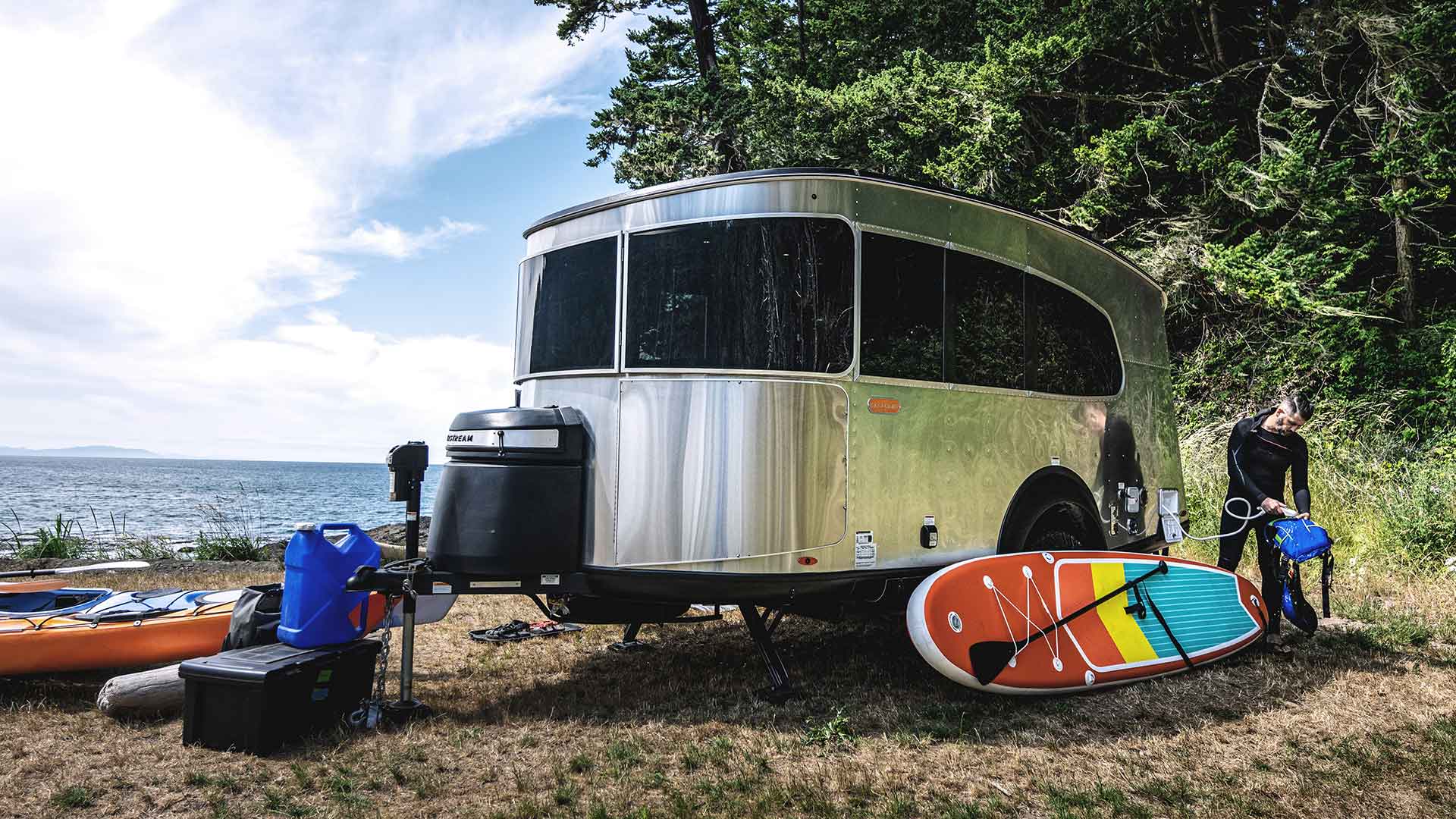A shiny camper trailer by a lakeside, surrounded by trees, with a paddleboard, kayak, and camping gear on the grassy ground.