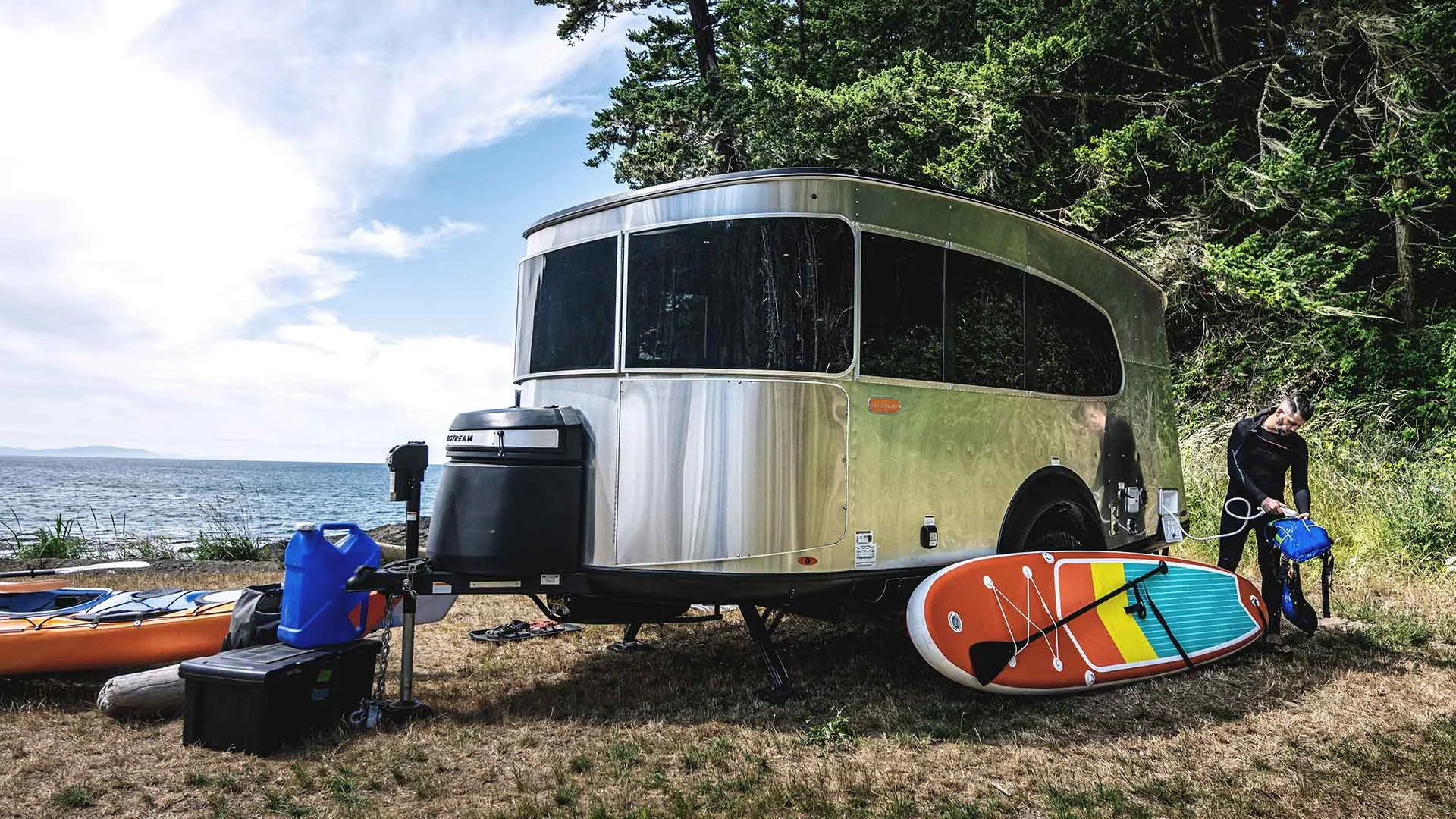 Airstream trailer by a lakeside with paddleboards and kayaks on the grass, surrounded by trees and cloudy sky.