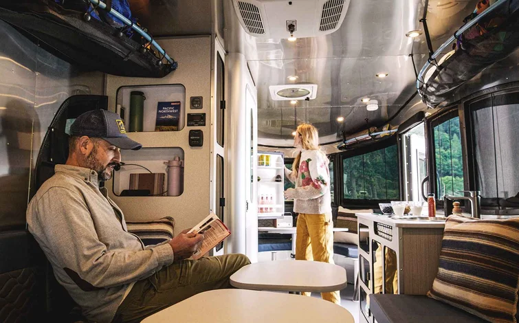 Man reading and woman standing near refrigerator inside a modern RV, with sleek interior design and large windows overlooking greenery.