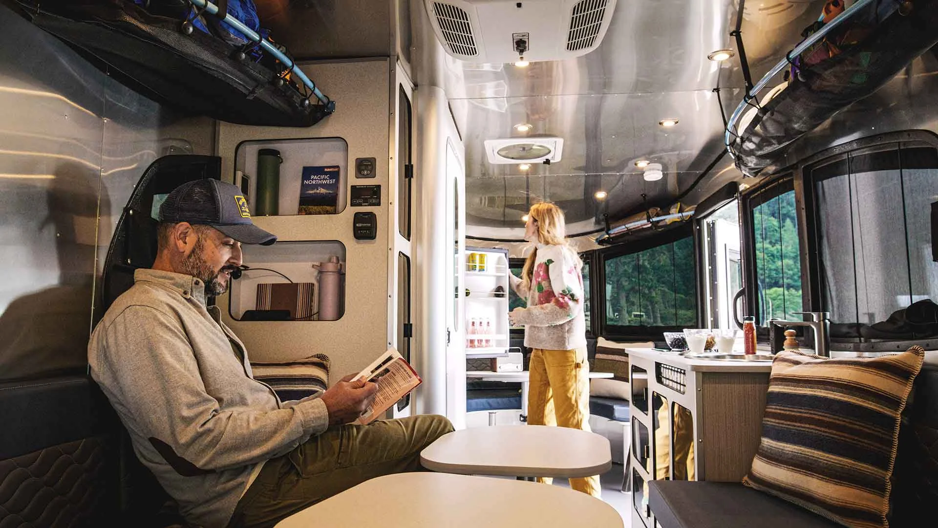 Man reading in a modern RV interior while a woman checks the fridge. Bright, organized space with large windows showing greenery outside.