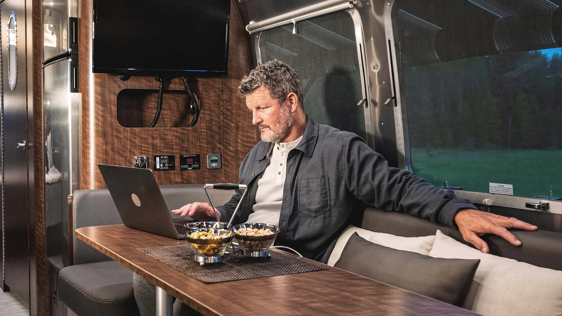 Man working on laptop inside an RV with wooden interior, seated at a table with snacks. Large window shows greenery outside.