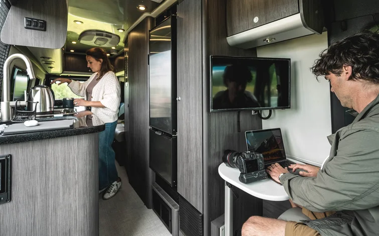 A man works on a laptop at a small table with a camera nearby while a woman prepares a drink in the kitchen area of a modern camper van.