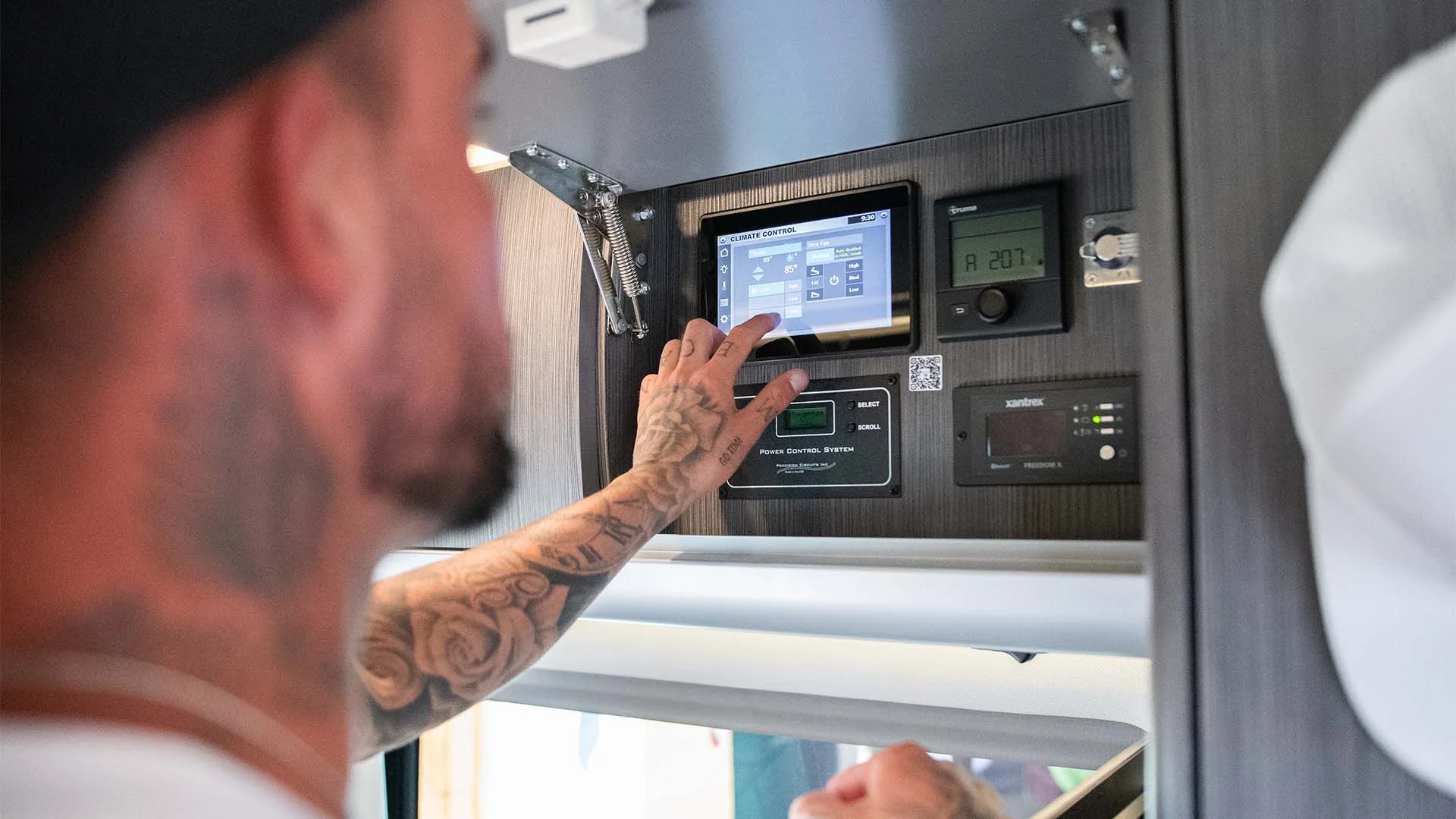 A person with tattoos adjusts a digital control panel inside a vehicle, surrounded by various electronic displays and buttons.