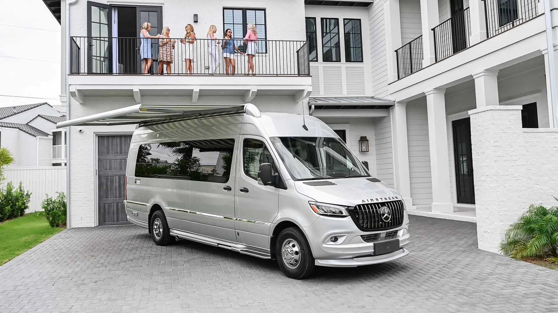 Silver van with an extended awning parked in front of a modern, white house. Group of people stand on the balcony above.