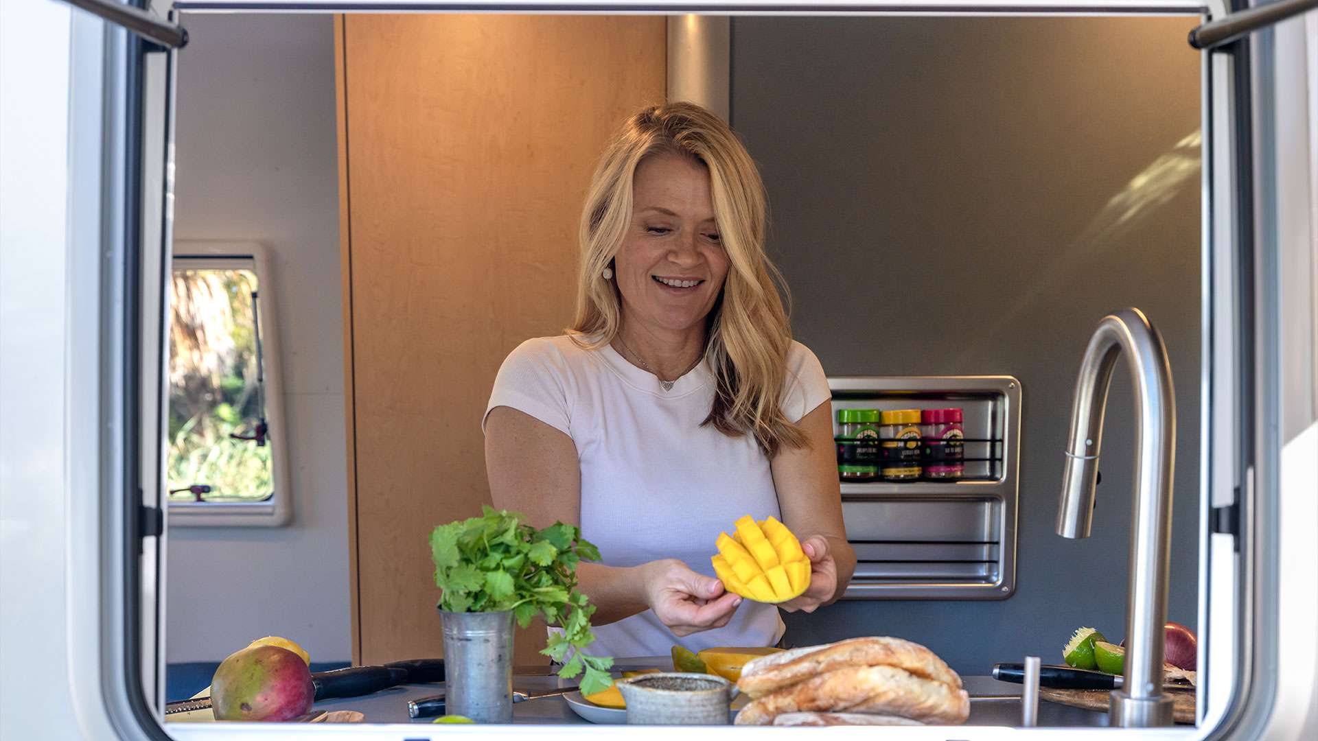 A woman in a white shirt prepares a sliced mango in a cozy kitchen, with herbs, bread, and spices on the counter.