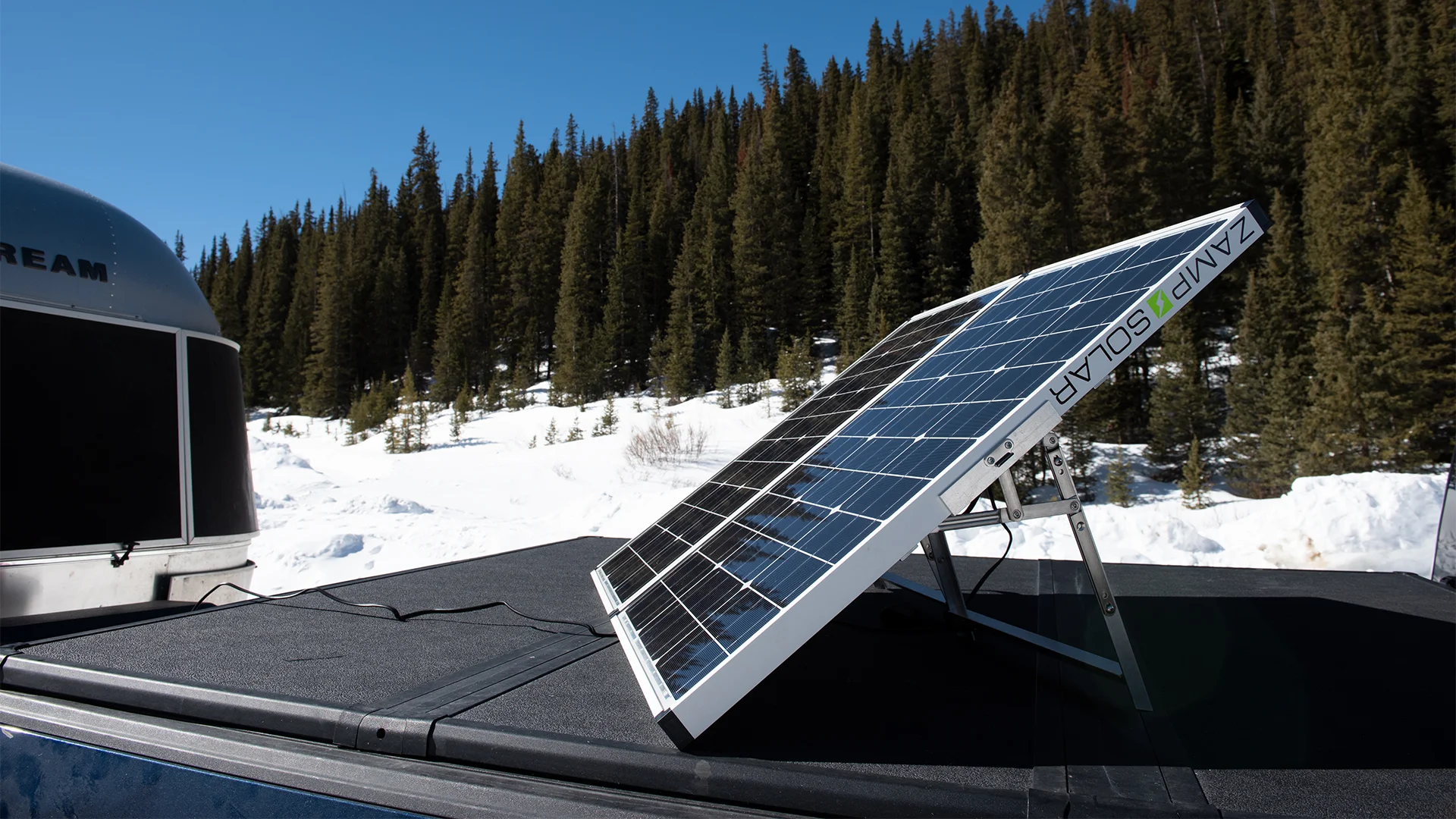 A portable solar panel is set up on a vehicle roof in a snowy, forested mountain area under a clear blue sky. Pine trees and part of a silver trailer are visible in the background.