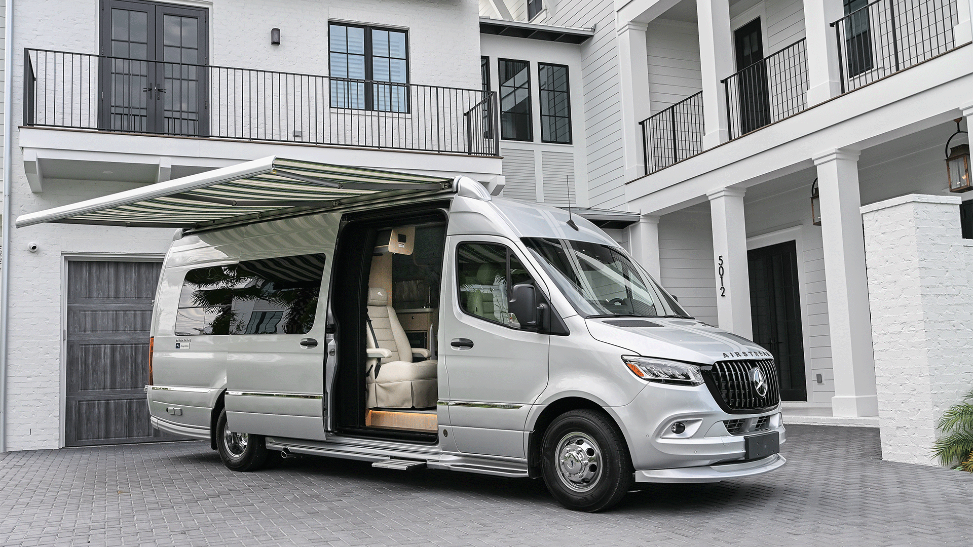 A silver luxury camper van with an extended awning is parked in front of a modern, white-brick house with large windows and a balcony.