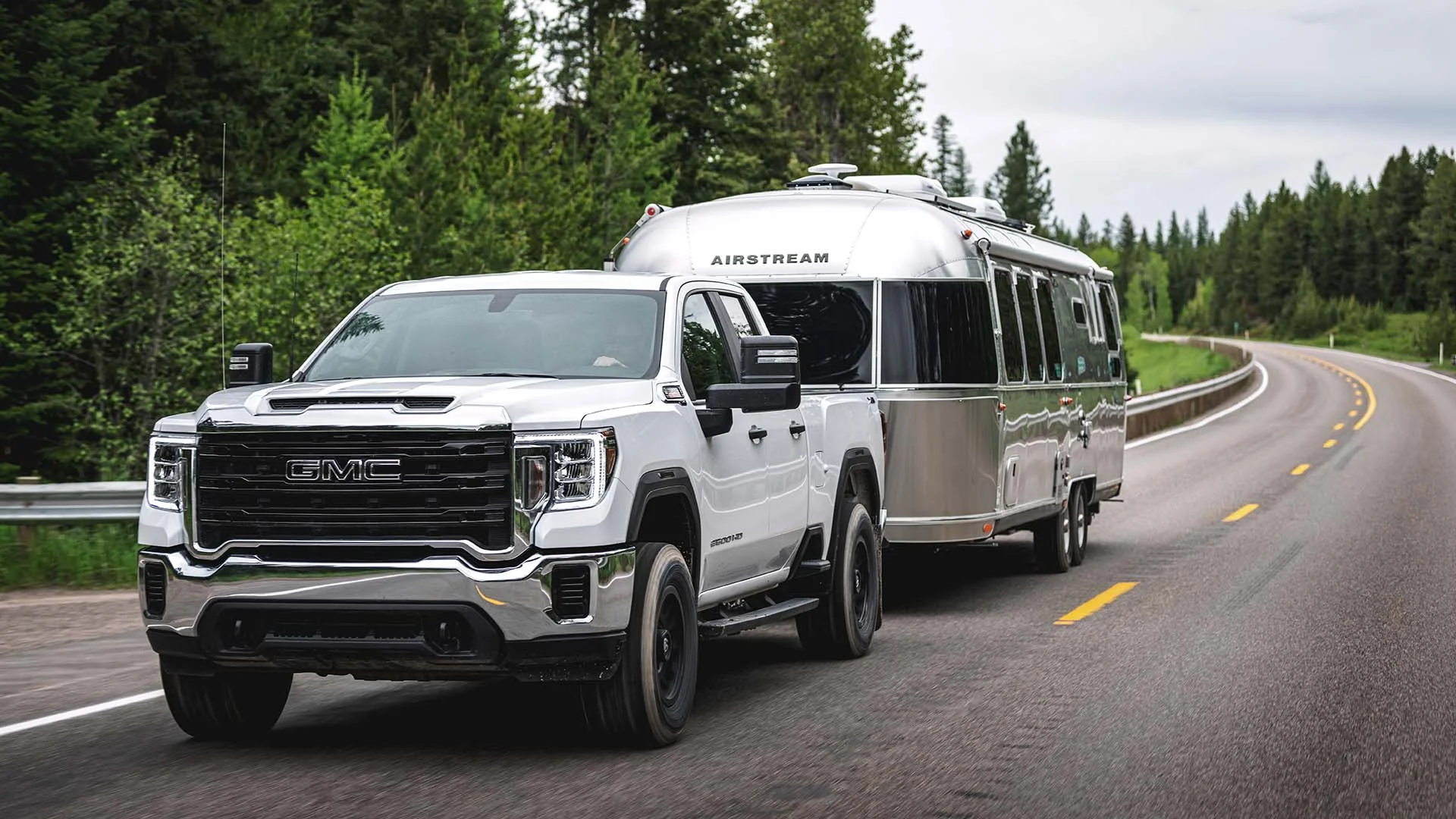 White GMC truck towing a silver Airstream trailer on a winding road surrounded by lush green trees.