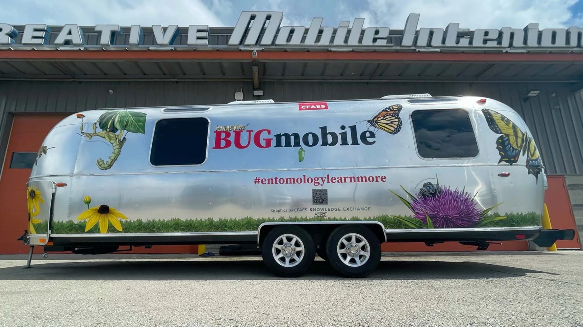 Silver trailer with "BUGmobile" text, butterfly and insect graphics, parked under "Mobile" sign. Has hashtag #entomologylearnmore.