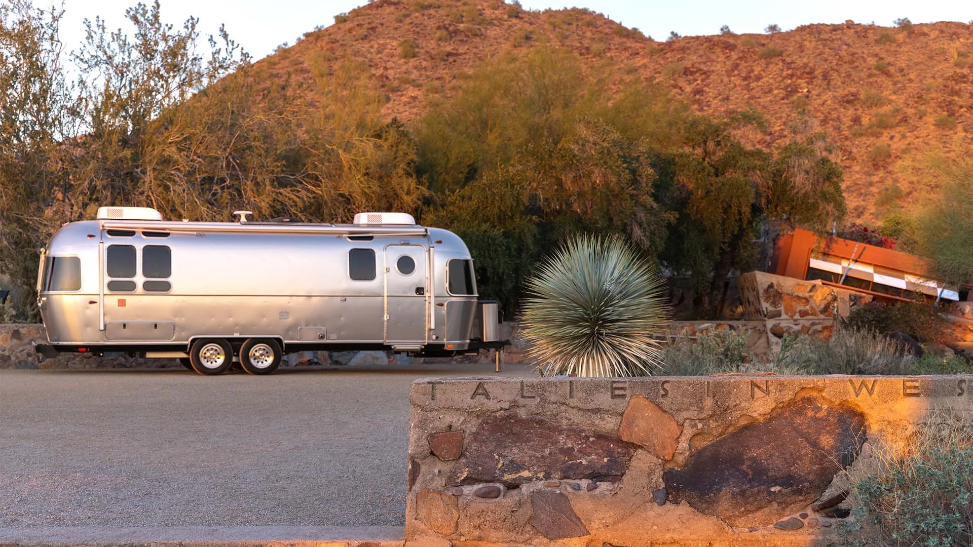 A silver Airstream trailer parked on a desert road beside a rocky sign. Desert hills and vegetation are illuminated by golden sunlight.