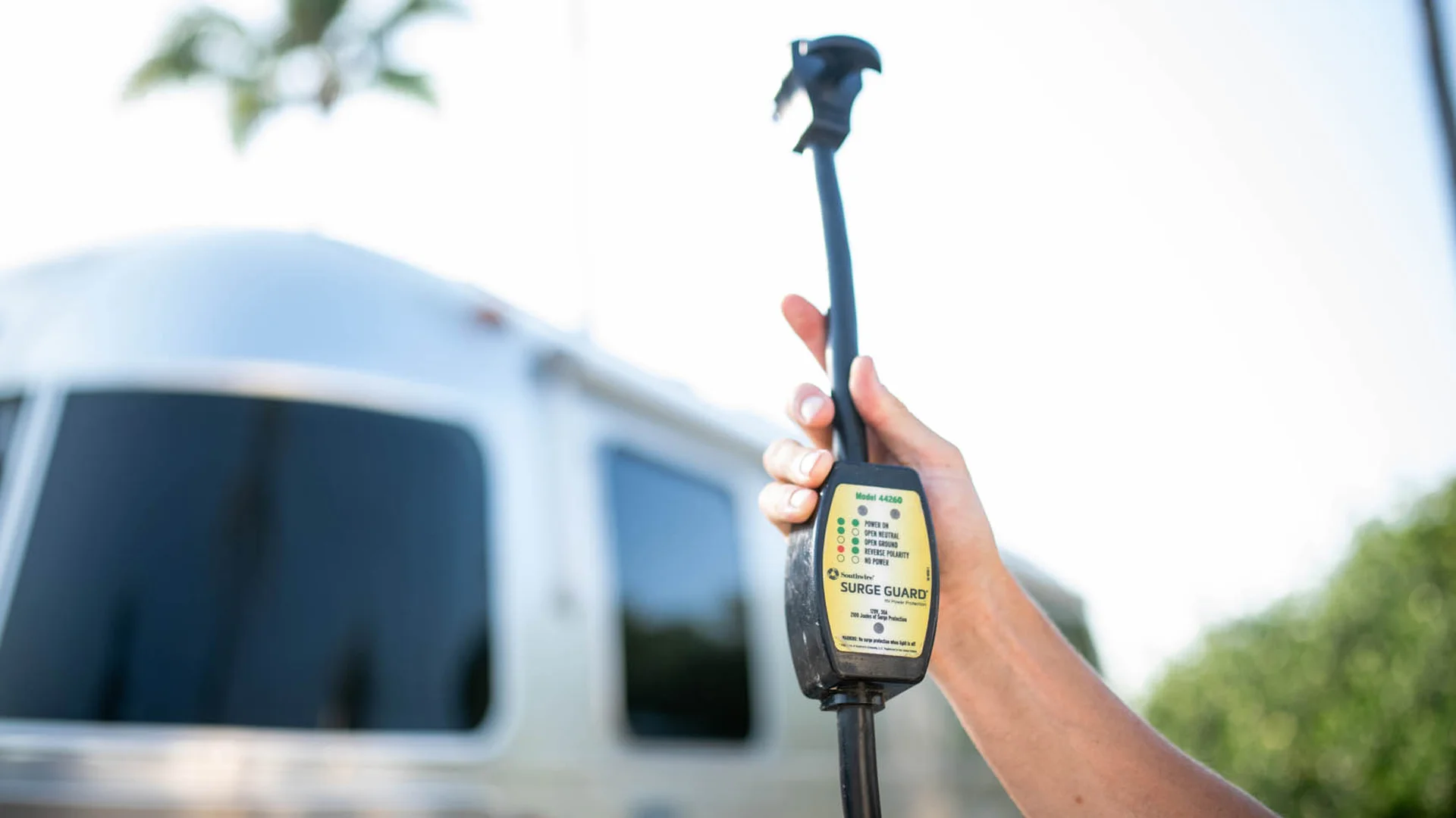 A person holds up a black RV surge guard power adapter with indicator lights, in front of a silver RV with dark tinted windows and a blurred green outdoor background.