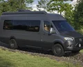 A dark gray camper van drives on a rocky path through a grassy, wooded area under a partly cloudy sky. The vehicle has large windows and off-road tires, surrounded by green trees and shrubbery.