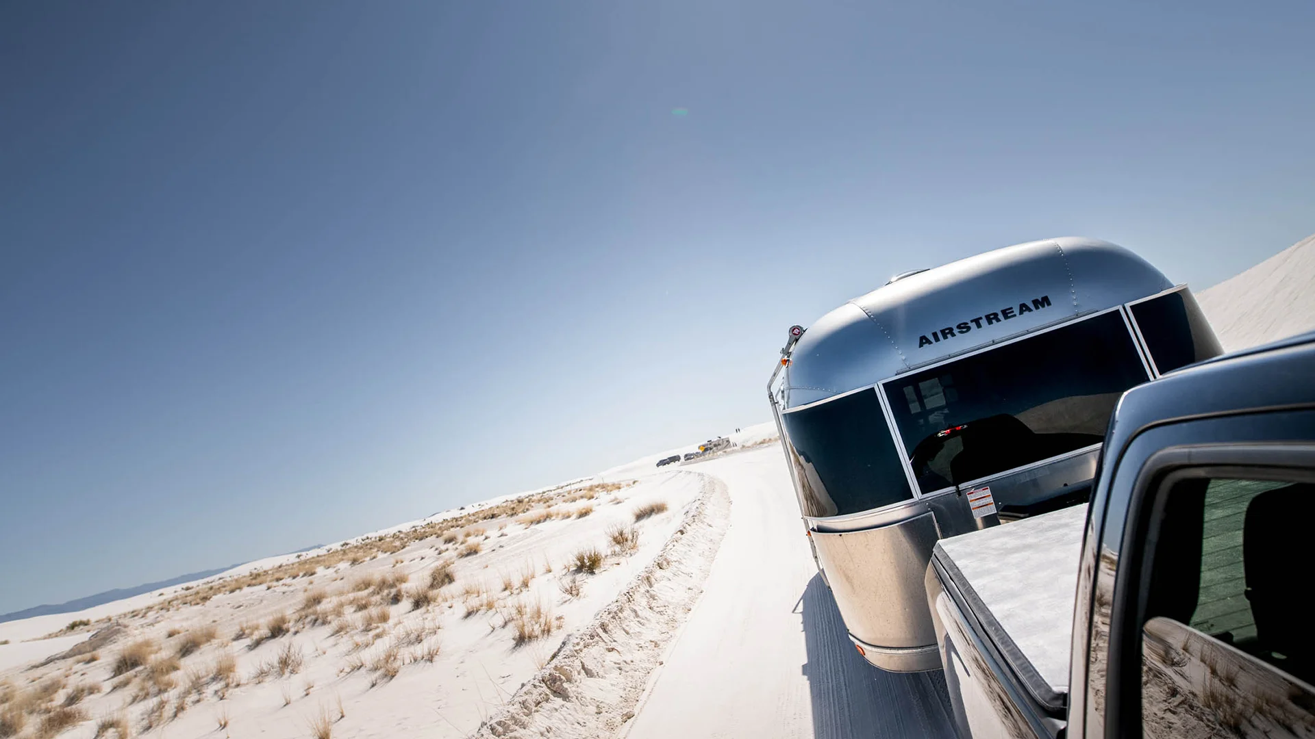 A silver Airstream trailer is being towed by a vehicle on a sandy desert road, under a clear blue sky. Sparse vegetation is visible along the road.