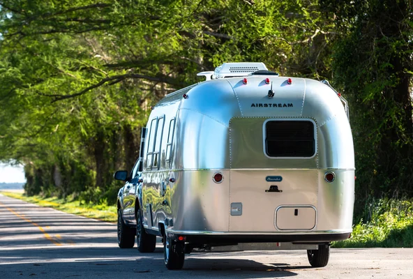 Silver Airstream trailer towed by a vehicle on a scenic road, with green trees lining the side under a clear blue sky.