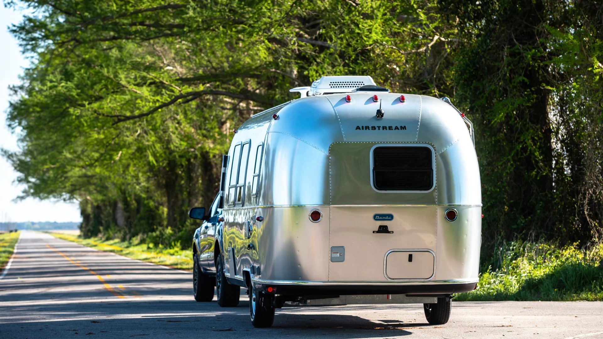 Silver Airstream trailer towed by a vehicle on a scenic road, with green trees lining the side under a clear blue sky.
